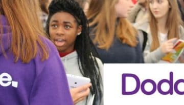 BAM Agency A woman wearing a purple hoodie with Doddle printed on the back engages with a diverse group during the exciting Doddle launch. In the background, someone holds a pamphlet, highlighting the youth marketing campaign, alongside the prominently displayed Doddle logo. BAM Agency