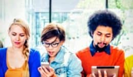 BAM Agency A diverse group of seven people stands in a row, engrossed in their phones and tablets—a snapshot of modern social media habits. Theyre in a bright room with large windows and greenery outside, each person expressing a mix of styles and emotions. BAM Agency