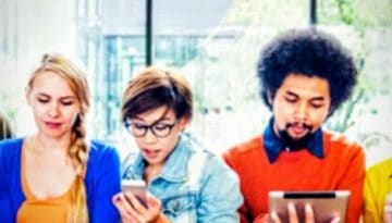 BAM Agency A diverse group of seven people stands in a row, engrossed in their phones and tablets—a snapshot of modern social media habits. Theyre in a bright room with large windows and greenery outside, each person expressing a mix of styles and emotions. BAM Agency