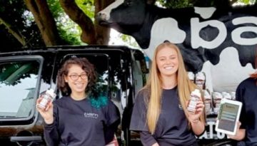 BAM Agency Three people stand in front of a truck topped with a cow model, each holding drinks and tablets. Wearing matching shirts with the Bam Life University logo, they proudly showcase their milkshake makers. The truck bed is filled with product bottles against a backdrop of trees. BAM Agency
