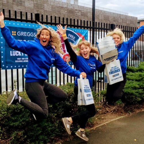 Three women in blue hoodies joyfully jump in the air, holding bags and boxes that have Greggs visible on them. Behind them is a black fence with banners advertising Greggs offers. Bushes and a road are in the background. BAM Agency