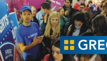 BAM Agency During a bustling Greggs promotional event, a crowd swarms around the booth as part of their Freshers tour. A staff member in a blue Greggs shirt enthusiastically engages with attendees, while the iconic Greggs logo is prominently displayed on the booth. BAM Agency