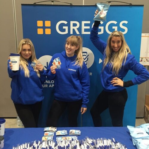 Three women wearing blue Greggs hoodies pose in front of a Greggs display table and banner. They hold promotional items, including a cup, packet, and card, standing in a cheerful, engaging manner. BAM Agency
