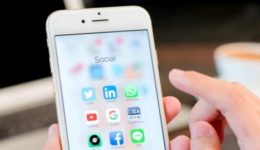 BAM Agency A person in 2017 holds a smartphone showcasing a menu of social media apps like Twitter, LinkedIn, WhatsApp, and Instagram. In the backdrop, a blurred laptop and cup hint at a setting where social media plays a key role. BAM Agency