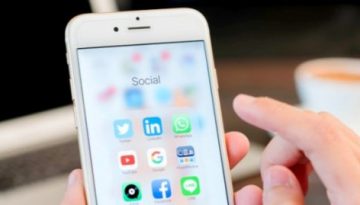 BAM Agency A person in 2017 holds a smartphone showcasing a menu of social media apps like Twitter, LinkedIn, WhatsApp, and Instagram. In the backdrop, a blurred laptop and cup hint at a setting where social media plays a key role. BAM Agency