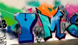 BAM Agency A vibrant graffiti mural on a white brick wall celebrates the YMS London 2018 event highlights. The artwork features animated letters YMS in bold colors: blue, orange, and pink, with dynamic designs and playful faces. The left side includes cartoon-style doodles of a computer, cityscape, and clouds. BAM Agency