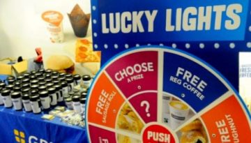 BAM Agency A display booth adorned with blue Greggs Rewards banners features a Lucky Lights game wheel and enticing promotional offers for freebies. Stacked boxes, coffee cups, and a sign urging app downloads beckon, making it a vibrant stop on the youth marketing journey. BAM Agency