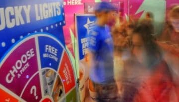 BAM Agency People gather around a colorful booth with a spinning wheel titled Lucky Lights, part of a vibrant marketing campaign. The wheel offers options like Choose, Free, and numbers. The scene is lively, with attendees appearing in motion, creating a blurred effect. BAM Agency