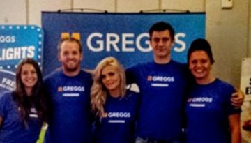 BAM Agency A group of six people wearing blue Greggs shirts stands in front of Greggs-themed posters and a spinning wheel display, smiling for the picture. It appears to be a lively Freshers event indoors, possibly managed by an enthusiastic Event Manager. BAM Agency