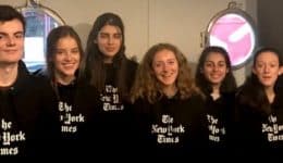 BAM Agency A group of eight brand ambassadors stands in line, each donning a black hoodie emblazoned with The New York Times logo. Indoors, against a backdrop of circular windows and a black wall featuring the iconic logo, they all smile brightly, embodying the spirit of this campaign. BAM Agency