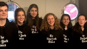 BAM Agency A group of eight brand ambassadors stands in line, each donning a black hoodie emblazoned with The New York Times logo. Indoors, against a backdrop of circular windows and a black wall featuring the iconic logo, they all smile brightly, embodying the spirit of this campaign. BAM Agency