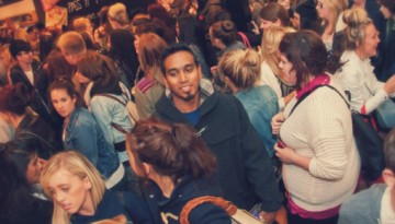 BAM Agency A crowded scene with people of various ages and ethnicities closely packed together at a vibrant UK student shopping event. Many engage in conversation or look around, the atmosphere lively with dim, warm lighting, suggesting a bustling BAM social gathering. BAM Agency