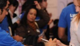 BAM Agency A person in a blue Lycamobile shirt hands a paper to a smiling student in a white shirt at a crowded indoor event, capturing the student appeal. Other attendees mingle in the background. BAM Agency