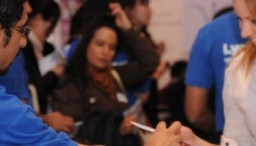 BAM Agency A person in a blue Lycamobile shirt hands a paper to a smiling student in a white shirt at a crowded indoor event, capturing the student appeal. Other attendees mingle in the background. BAM Agency