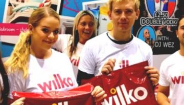 BAM Agency Four people wearing white Wilko t-shirts smile at the camera, holding Wilko bags. A large Wilko logo is visible in the background, capturing a vibrant moment of University Freshers engaged in dynamic youth marketing. BAM Agency