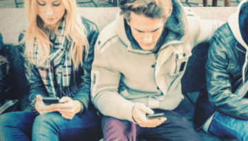 BAM Agency Four millennials sit on a stone bench, each engrossed in their smartphones. Dressed in casual winter attire with jackets and scarves, they exemplify the seamless integration of mobile marketing into social settings. BAM Agency