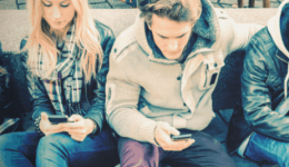 BAM Agency Four young adults, possibly students, sit on an outdoor bench, each focused on their smartphone and engaging with social media. Dressed in casual jackets and jeans, they create a relaxed atmosphere that suggests a casual hangout. BAM Agency
