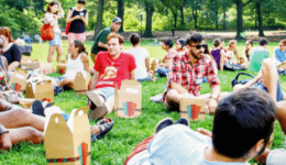 BAM Agency A diverse group of students is sitting on the grass in a park, enjoying a summer picnic. They are gathered in small groups, engaged in lively conversations and sharing food. Trees and greenery frame the background, creating a relaxed and social atmosphere. BAM Agency