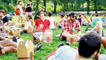 BAM Agency A diverse group of students is sitting on the grass in a park, enjoying a summer picnic. They are gathered in small groups, engaged in lively conversations and sharing food. Trees and greenery frame the background, creating a relaxed and social atmosphere. BAM Agency