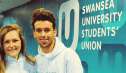 BAM Agency Three people in white hoodies stand smiling before a wall that reads Swansea University Students Union and its Welsh translation. The background boasts a modern, colorful design, showcasing the vibrant spirit of Student Ambassadors representing their university brand. BAM Agency