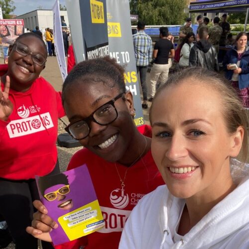 Three people are taking a selfie outdoors. Two wear red shirts with PROTONS emblazoned on them, while the third holds a colorful booklet. In the background, informational banners catch the eye alongside groups that appear to echo National Expresss vibrant community events. BAM Agency