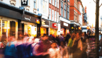 BAM Agency A bustling city street with blurred motion capturing countless pedestrians, reflecting both in-store shopping energy and students shopping habits. Storefronts with bright signs line the sides, while trees dot the sidewalk, conveying a fast-paced urban life. BAM Agency