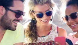 BAM Agency Three people wearing sunglasses are sitting together outside, smiling, and looking at a smartphone. Their relaxed and happy demeanor under the sunny sky perfectly captures the essence of a vibrant youth marketing strategy. BAM Agency