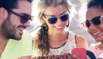 BAM Agency Three people wearing sunglasses are sitting together outside, smiling, and looking at a smartphone. Their relaxed and happy demeanor under the sunny sky perfectly captures the essence of a vibrant youth marketing strategy. BAM Agency