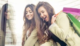 BAM Agency Two women with long hair smile while shopping, holding colorful bags. They are looking at a store window display in an urban setting, reflecting excitement and joy—a perfect scene for student marketing to capture vibrant energy and youthful enthusiasm. BAM Agency