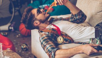 BAM Agency A man wearing a plaid shirt and sunglasses lounges on a couch, proudly sporting a 1st medal around his neck. Amidst the clutter of basketballs and scattered papers, he indulges in pizza and grips a game controller, embodying youthful trends shaped by the cozy behaviours of the COVID-19 era. BAM Agency