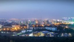 BAM Agency Panoramic view of a cityscape at dusk in the heart of Kazakhstan, featuring tall, illuminated skyscrapers on the left and a brightly lit cluster of buildings on the right against a twilight sky. Streets and city lights spread across the horizon. BAM Agency