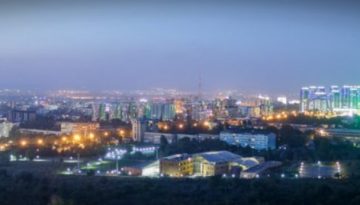 BAM Agency Panoramic view of a cityscape at dusk in the heart of Kazakhstan, featuring tall, illuminated skyscrapers on the left and a brightly lit cluster of buildings on the right against a twilight sky. Streets and city lights spread across the horizon. BAM Agency