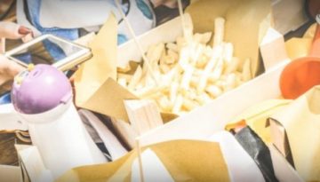 BAM Agency Millennials gather around a table at the restaurant, surrounded by fast food delights like fries, burgers, and drinks. Several hands clutch smartphones, capturing moments of connection in a modern social scene. BAM Agency