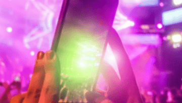 BAM Agency A smartphone held up in a colorful, crowded concert setting captures vibrant lights and stage effects. The scene buzzes with energy, featuring purple, pink, and green hues. Embracing trends from 2019, the blurred lights add a touch of nostalgia to the electric atmosphere. BAM Agency