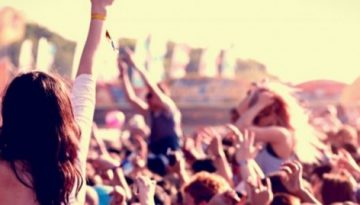BAM Agency A large crowd at an outdoor music festival dances and cheers, fully immersed in the festival experience. In the foreground, a person with arms raised soaks up the vibrant atmosphere. Colorful tents and stages brighten the background under a sunny sky. BAM Agency