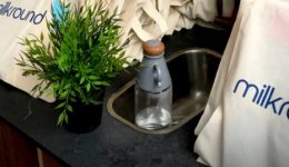 BAM Agency A countertop displays multiple canvas bags with the milkround logo, featuring a stylized milk bottle, as if ready for a university tour. Nearby, a small green plant and a transparent bottle with a wooden cap sit peacefully next to the sink. BAM Agency