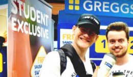 BAM Agency Two people are smiling in front of a Greggs promotion booth with Lucky Locker signage, capturing the essence of experiential marketing. Theyre holding drinks and wearing backpacks as they engage with the youth market. A third person is partially visible, facing a Student Exclusive banner. BAM Agency