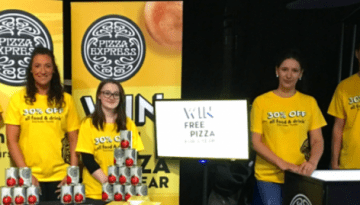 BAM Agency A group of seven people stands behind tables displaying cans, clad in yellow T-shirts with black text. A sign reads WIN FREE PIZZA, as PizzaExpress branding decorates the backdrop. Their presence at the event is a journey into captivating the vibrant student market. BAM Agency