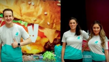 BAM Agency Four university students in Deliveroo t-shirts stand smiling in front of branded banners. The backdrop features a large, colorful image of a burger alongside the vibrant Deliveroo logo, capturing the essence of youthful energy and excitement in todays market. BAM Agency