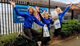 BAM Agency Three people in blue Greggs shirts are joyfully jumping in front of a fence, targeting the student market. They hold blue bags that say Today You Get The Freebies, while a Greggs banner is visible in the background. BAM Agency