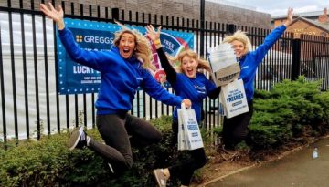 BAM Agency Three people in blue Greggs shirts are joyfully jumping in front of a fence, targeting the student market. They hold blue bags that say Today You Get The Freebies, while a Greggs banner is visible in the background. BAM Agency