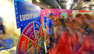 BAM Agency A bustling event scene unfolds with a Greggs Lucky Lights game wheel at its center, attracting a lively student market. The blurred motion captures the dynamic energy, while bright promotional signs and booths in the background encourage attendees to reconnect and engage safely. BAM Agency