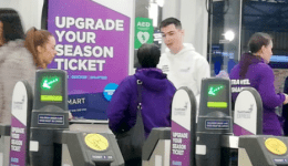 BAM Agency People walking through ticket barriers at a bustling transit station. Staff in purple jackets assist passengers as a Transpennine campaign encourages upgrading to a season ticket with a Smart Card. The scene is lively, filled with passengers and helpful staff. BAM Agency
