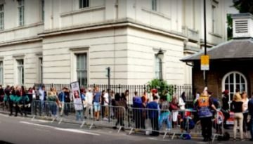 BAM Agency A long line of people eagerly awaits on the sidewalk outside a large, historic white building, hinting at an exciting event. Metal barricades divide the street, and behind them, a columned structure peeks through the trees like something out of a dead-or-alive mystery. BAM Agency