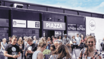 BAM Agency A bustling outdoor scene with a crowd gathered in front of modern storefronts, including Starbucks and Piazza. Among the lively atmosphere, Southampton Students Union students chat animatedly under the clear blue sky, adding to the vibrant energy of the day. BAM Agency
