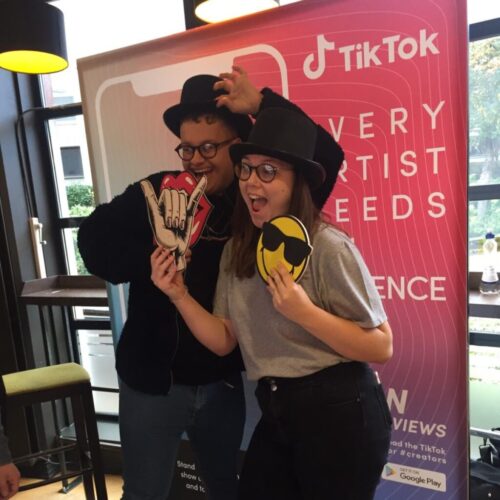 Two people wearing hats pose playfully with emoji and gesture props in front of a TikTok banner. One holds a rock hand gesture and lips, the other holds a smiley face with sunglasses. They stand in a room with tables and chairs. BAM Agency