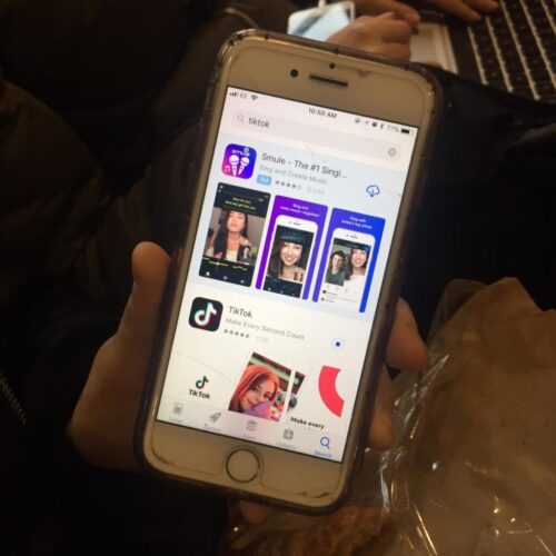 A person holding a smartphone with the App Store open, showing a search for TikTok. The TikTok app and other related apps are displayed on the screen. A laptop is partially visible in the background. BAM Agency