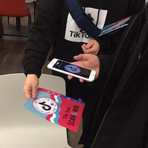 Two people holding a TikTok promotional flyer and a smartphone. One person uses the phone, opened to a scanning app, possibly engaging with the Scan & Win feature on the flyer. They are in a room with chairs and a table. BAM Agency