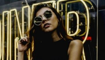 BAM Agency A woman in sunglasses stands in front of a reflective sign with the word DINER in large yellow neon letters. Sporting a sleeveless black top, she runs one hand through her hair, embodying the modern, urban vibe often seen in Instagram Influencer posts. BAM Agency