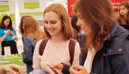 BAM Agency At a bustling job fair, people engage around vibrant stalls. A person in a blue shirt hands out information to attentive attendees clutching marketing pamphlets. Bright banners promoting local brands surround the area, adding to the lively atmosphere. BAM Agency